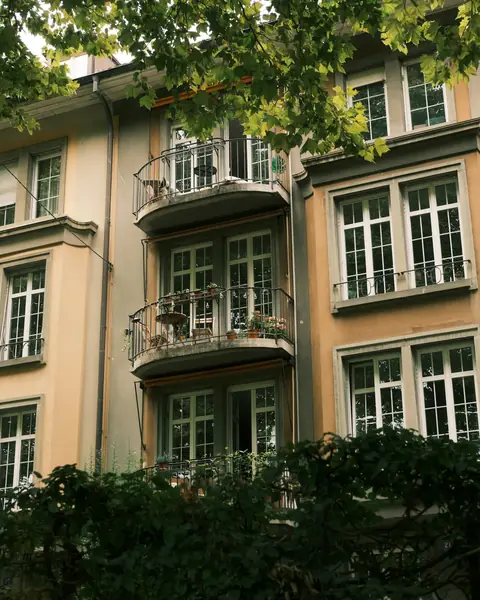 Ağaçların arasında, balkonlu ve yüksek pencereli bir apartman cephesi